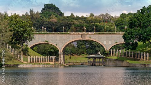Stone arch bridge over Putrajaya Lake, featuring a charming gazebo, lush green hills, and decorative lampposts. Located in Putrajaya, Kuala Lumpur, Malaysia.