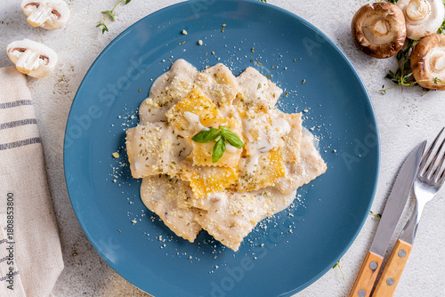 a plate of ravioli, filled with a butternut squash mixture, served in a creamy white sauce. It is garnished with grated Parmesan cheese, dried herbs, and a fresh basil leaf, with fresh mushroom