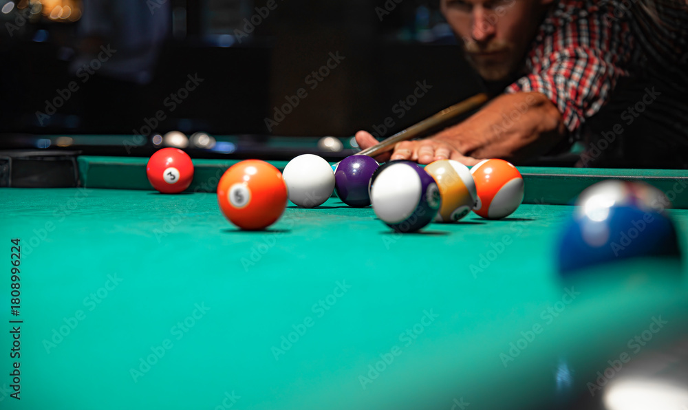 Naklejka premium A close-up view of colorful billiard balls scattered on a green felt table while a focused man in a plaid shirt aims his cue stick in the blurred background.