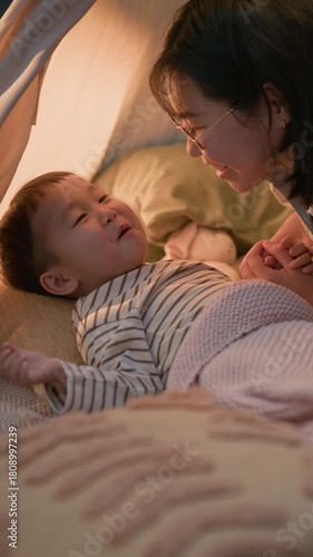 Vertical shot of caring Asian mother massaging little son belly and kissing him gently on cheek while calming him down before night sleep in soft bed, creating relaxing atmosphere