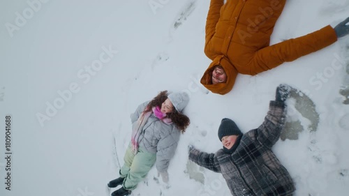From above shot of parents and child lying on snow side by side with arms spread out in open clearing surrounded by winter landscape