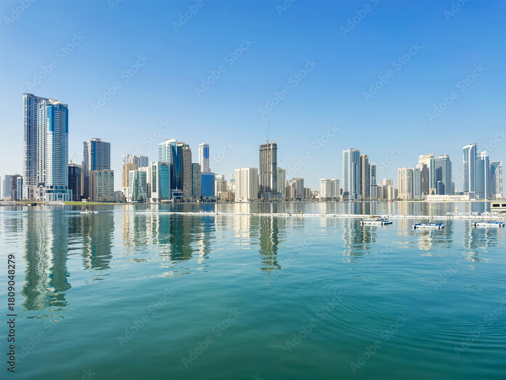 Naklejka premium View from Al Majaz Corniche in Sharjah, UAE, featuring a calm waterfront and the modern skyline in the distance, a clean cityscape with soft light, simple geometry, and a calming, balanced atmosphere.
