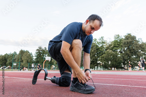Athlete with a disability, wearing a prosthetic running blade, pausing on the track to tie his shoelace on a sneaker. Motivation and persistence concept.