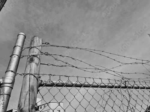 Black and white close-up of barbed wire attached to a wooden and metal post, running above a wire fence, with a cloudy sky in the background.
