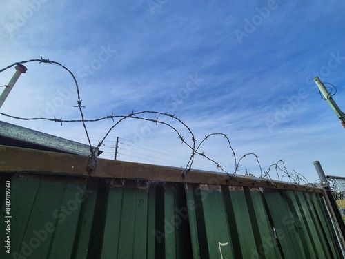 Close-up of barbed wire coiled around the top edge of a fence, against a whispy cloudy sky.
