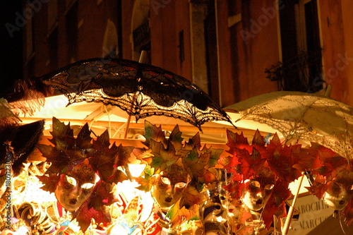 A glowing Venetian mask display at night, showcasing ornate leaf-inspired designs and lace parasols. A vibrant glimpse into Venice’s carnival artistry and festive charm.