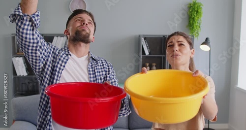 Couple catching ceiling water leak. They use buckets indoors to catch dripping water from a damaged roof, showing home stress and urgent repair. Leakage and urgent home water damage.