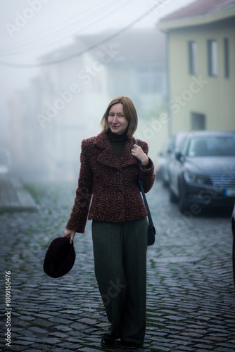 Woman standing on a foggy cobblestone street, holding a hat, dressed in a textured brown jacket and green trousers, with soft morning mist wrapping the quiet neighborhood.