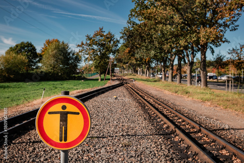 A striking yellow and red warning sign prohibiting access stands prominently on the narrow-gauge railway tracks on Rügen, framed by autumn trees and a blue sky.