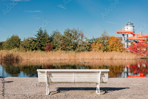A white bench overlooks a pond reflecting autumn trees and the tower and orange water slides of a modern spa or leisure facility in the background.