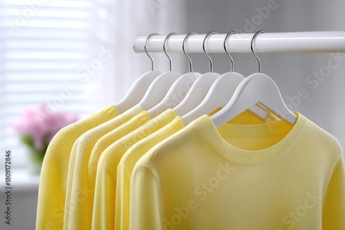 Wallpaper Mural Yellow Sweaters on Hangers: A row of neatly arranged yellow sweaters hang on sleek, white hangers against a soft, blurred backdrop of a window and a hint of a flower vase. Torontodigital.ca