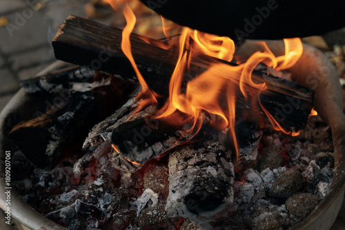A close-up (macro) of an open fire: a bright orange flame and smoldering red embers burning in a special metal tray. Concept: the element of fire, warmth, combustion, outdoor cooking.