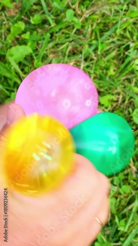Hands hold and press green, pink and yellow slime balls on bright grass, demonstrating elastic movements, playful tactile movements and summer relaxation Playing with a toy colorful anti-stress