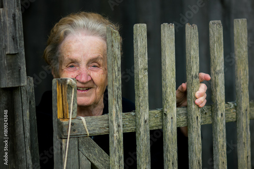 Smiling elderly woman peeks from behind an old wooden fence, her hand gently holding one of the weathered slats.