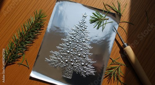 Silver foil decoration with pressed Christmas tree design on wooden surface. Pine needles and embossing stylus beside the artwork. Holiday tin foil embossed ornament, silver tree, rustic decor
