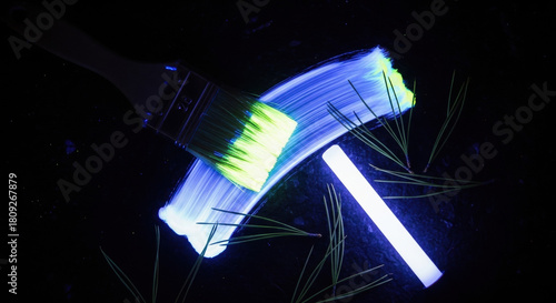 Paintbrush with glowing paint on bristles over dark surface. Pine needles and UV light wand arranged nearby. Phosphorescent paint brush stroke, glowing in dark party theme
