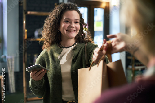 Wallpaper Mural Young adult Caucasian woman smiling while receiving shopping bag from another , holding smartphone in hand, standing indoors in modern retail environment Torontodigital.ca