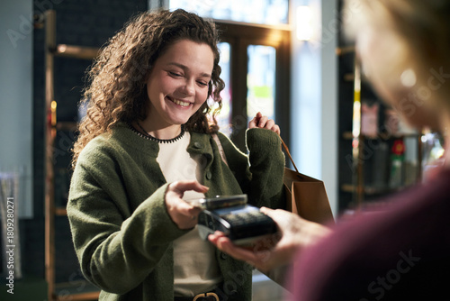 Young adult Caucasian woman smiling while making contactless payment with smartphone at checkout counter, holding shopping bag in modern retail store, interacting with cashier