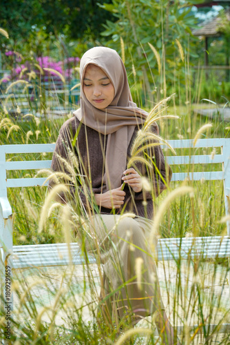 An Asian Muslim woman appears graceful and calm as she poses holding a weed on a park bench with a warm natural feel.