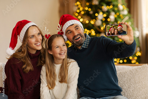 Cheerful family wearing Santa hats taking selfie with smartphone, sitting on couch in cozy living room, decorated with sparkling Christmas tree on background