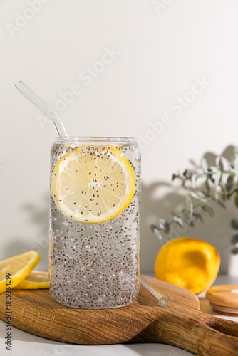 Refreshing lemon water with chia seeds in a clear glass with a glass straw on a wooden board in natural light.