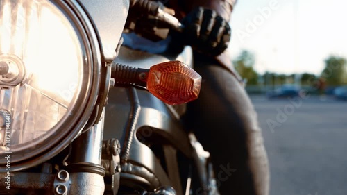 Close up shot of a silver retro motorcycle, with the biker resting one hand on the handle as the orange turn signal and circular headlight glow in the frame.