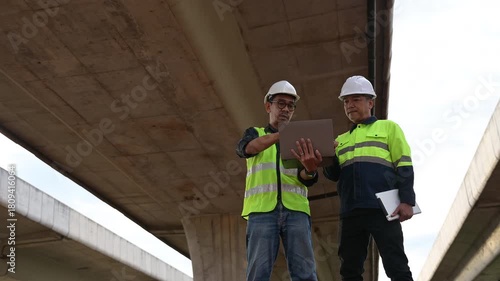 Bridge construction analysis: Two construction workers stand beneath a concrete bridge, meticulously examining blueprints, symbolizing collaboration and expertise in infrastructure projects.