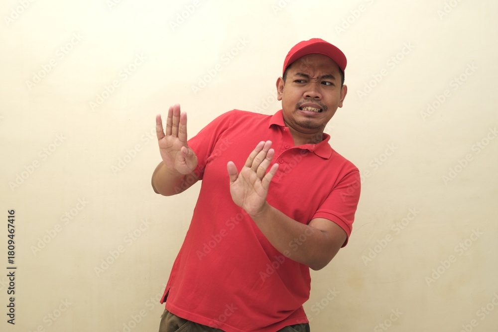 Fototapeta premium Southeast Asian man in red uniform leaning back with hands up in fear or rejection, isolated with copy space for advertisement use. 
