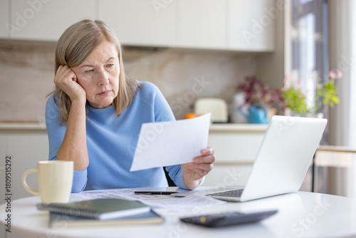 Senior woman feeling stressed and worried while reviewing financial documents and monthly bills, calculating her budget and dealing with debt issues in her home kitchen