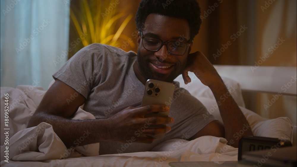 Obraz premium Happy amazed African American man male guy customer client lying down on bed at home late evening holding smartphone user stunned wow reading great news watching social media surprised amazed indoor