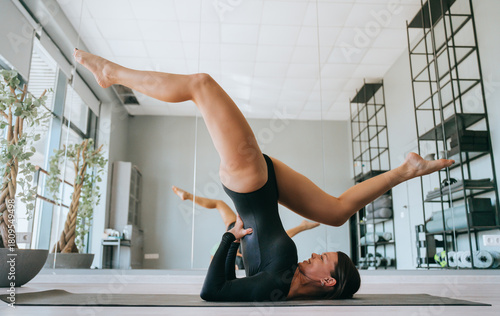 A woman in a black leotard performs a challenging yoga pose on a mat, gracefully balancing her legs above in a bright studio.