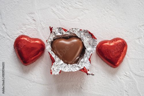 Heart-shaped chocolates wrapped in shiny red foil are arranged on a light-colored surface. One chocolate is open, showing a tasty chocolate heart inside.