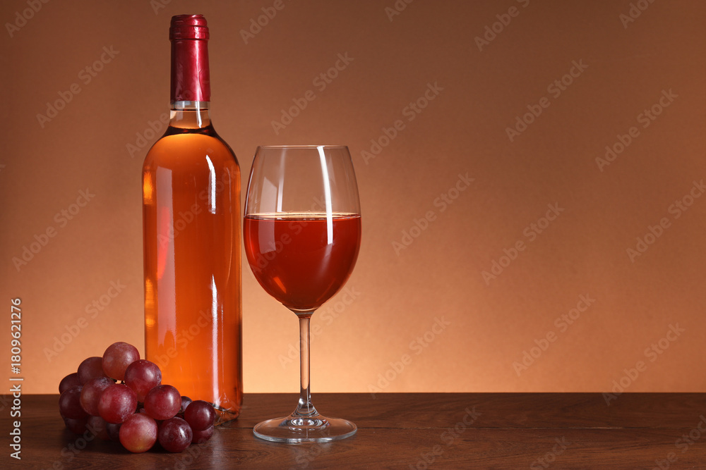 Fototapeta premium Rose wine in glass, bottle and grapes on wooden table against brown background, space for text