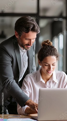 Manager Coaching Employee – Male manager leaning over desk to guide young employee on laptop work
