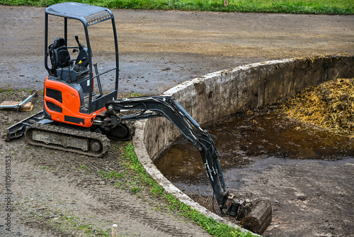 Mini excavator cleaning a circular manure pit on a farm, showing agricultural machinery at work in a rural environment