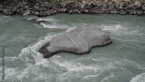 Large gray boulder partially submerged in rushing turquoise-white water and foam, geology concept.