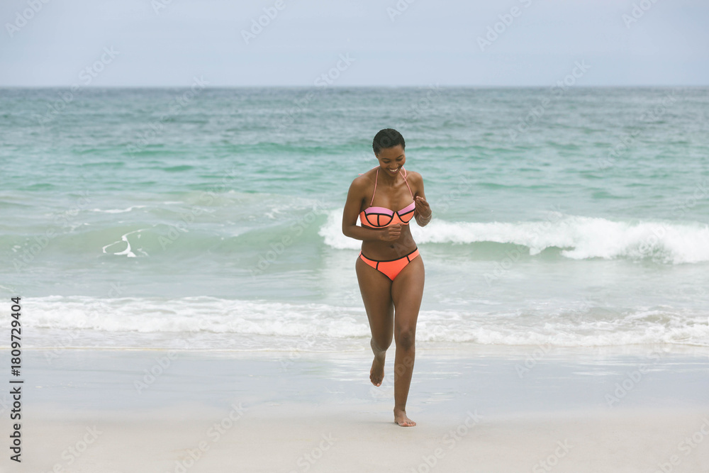 Naklejka premium African American woman walking from surf onto wet sand, adjusting bikini strap with horizon visible