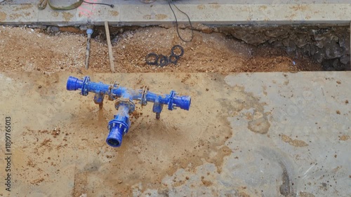 Top view of a muddy trench in a road construction site. A blue ductile iron water pipe T-joint is visible sitting in the mud and water, ready for installation. Construction tools, including a jackhamm