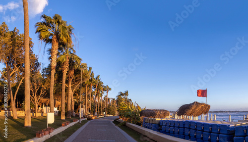 Fototapeta Naklejka Na Ścianę i Meble -  A tiled promenade stretches along the sea in Side, lined with palm trees and beaches. A red storm flag flies under a blue evening sky, showcasing the resort's quiet low season. Turkey.

