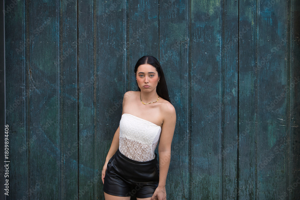 Fototapeta premium Young woman, attractive, brunette, blue eyes, Hispanic, wearing a white top and leather shorts, posing and looking at the camera leaning against a blue door in the background. Concept: model, beauty.