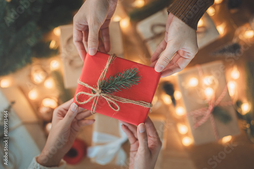 Christmas gift exchange with hands holding a beautifully wrapped red present decorated with natural twine and pine twig, symbolizing festive giving, warmth, celebration, and holiday tradition
