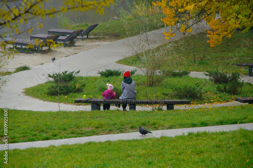 Autumn landscape. Young mother and daughter are sitting on a bench in an autumn park by the lake. Outdoor recreation.