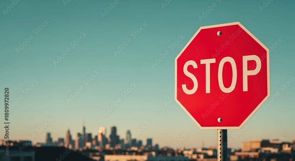 Obraz premium Red octagonal stop sign stands prominently against a clear blue sky, with a blurred city skyline in the background, emphasizing traffic regulations and safety awareness