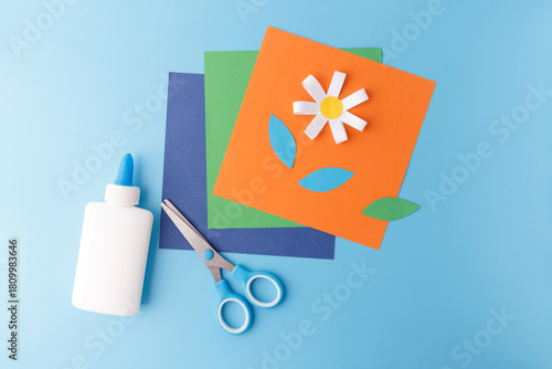 Paper flower craft with scissors, glue bottle and colored paper sheets on blue background. Flat lay composition. Kids craft and DIY project concept for design, poster, banner, greeting card 