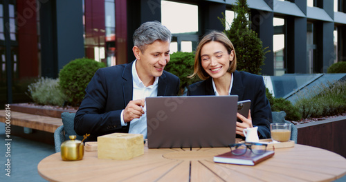 Business partners coworkers businessman and businesswoman working together in city with laptop writing notes in notebook. Man and woman in suits having fun, laughing, watching something funny on