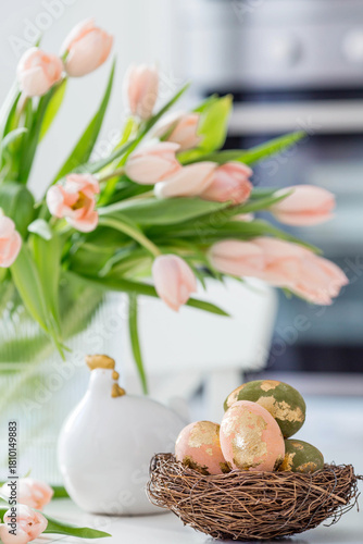 Beautiful Easter greeting card. A bouquet of delicate tulips, a chicken, and eggs with a golden pattern on a table in a white kitchen interior.