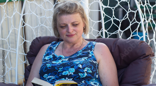 A beautiful woman is sitting in a rocking chair in the yard, reading a book.