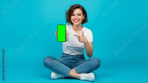 eye-level studio shot of a young, cheerful woman sitting cross-legged against a solid, bright cyan blue background.