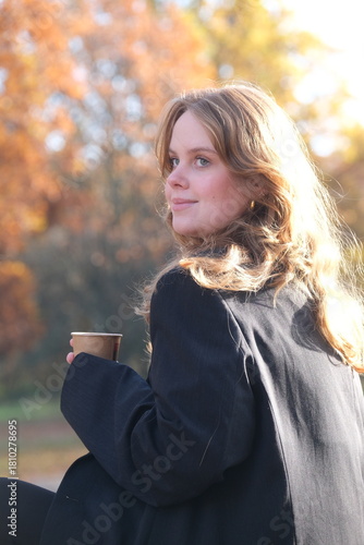 Konstfotografi Girl is sitting on a bench in city autumn park with paper cup of coffee