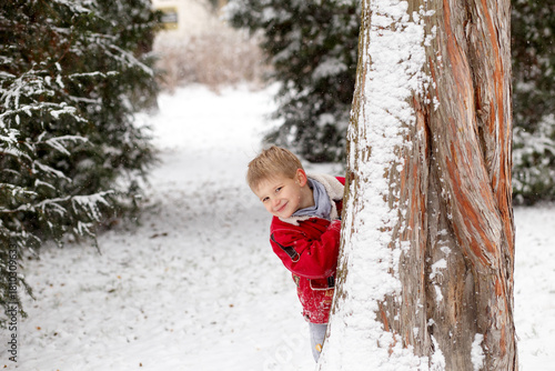 Tableau sur toile Smiling boy in a red winter jacket peeking from behind a snowy tree in a festive